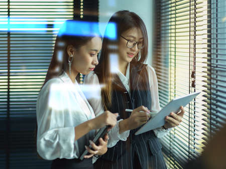 View through glass window of two female office workers consulting on their project while standing in officeの写真素材