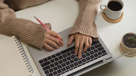 Top view of female hand typing on laptop keyboard on table with coffee cup and notebook
