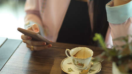 Close up view of female hand holding smartphone with tea cup on wooden table in cafeの写真素材