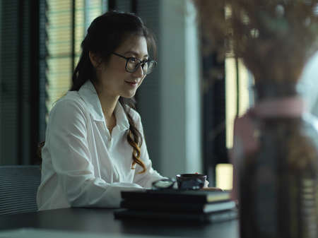 Side view of businesswoman working with computer device on office desk with plant vase and suppliesの写真素材
