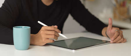 Close up view of a man using digital tablet with stylus on white table with coffee mugの写真素材