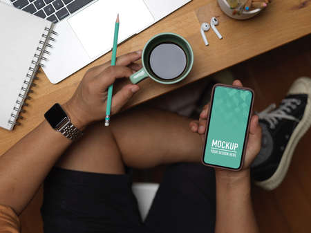 Overhead shot of male using smartphone and holding coffee cup while relaxing at workplace,