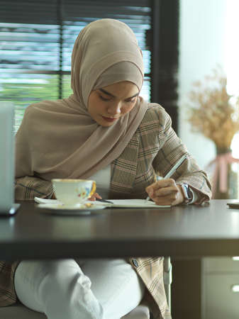 Portrait of young Asian muslim writing on notebook on office desk with cup and laptopの写真素材