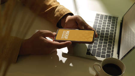 Side view of male hand holding smartphone on workspace with laptop and coffee cup