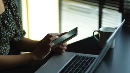Side view of female using smartphone while working with laptop on worktable in office roomの写真素材