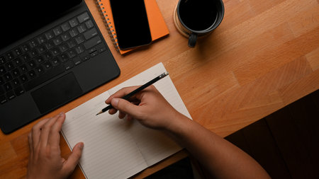 Top view of male hand writing on notebook on worktable with tablet, smartphone and coffee cup