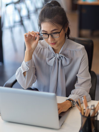 Portrait of businesswoman with eyeglasses working with laptop in office roomの写真素材