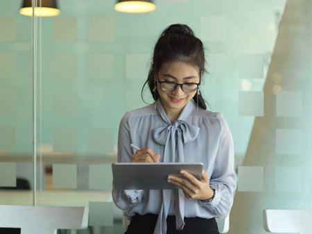 Portrait of businesswoman working with digital tablet while standing in glass partition office roomの写真素材