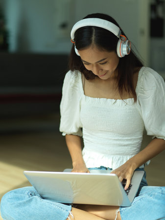 Portrait of female with headphone using laptop on her lap while crossed legs sitting on the floorの写真素材