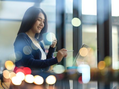View through glass wall with bokeh of female teenager doing assignment with digital tablet in cafeの写真素材