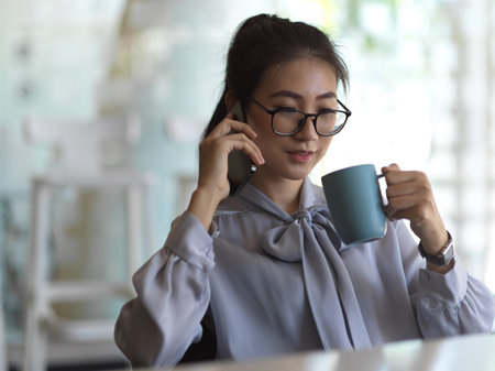 Portrait of businesswoman talking on the phone and drinking coffee while relaxing in office roomの写真素材