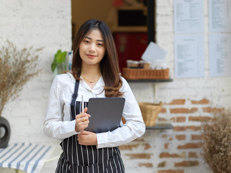 Portrait of young beautiful waitress holding digital tablet smiling to camera while standing in restaurantの写真素材