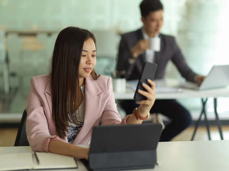 Portrait of businesswoman using smartphone while working with digital tablet in office roomの写真素材