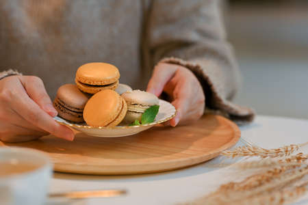Close up view of female holding a plate of French colourful macarons from wooden tray on coffee...