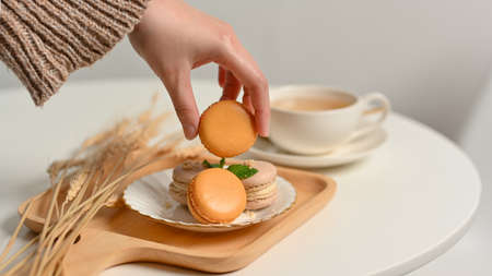 Close up view of female hand picking French macaron from a plate on coffee table with tae cup