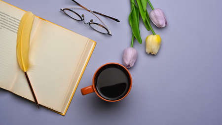 Top view of workspace with opened book, coffee up, eye glasses and tulips flower on purple table