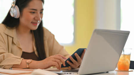 Close up view of female teenager with headphone using smartphone while working with laptop in cafe