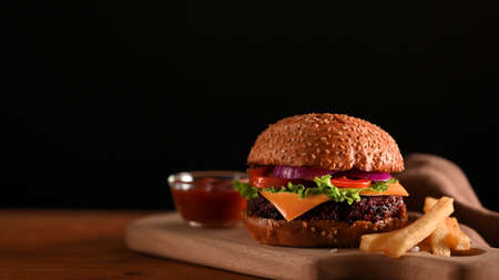 Close-up view of grilled beef burger served on wooden tray on the table with ketchup and black wall...