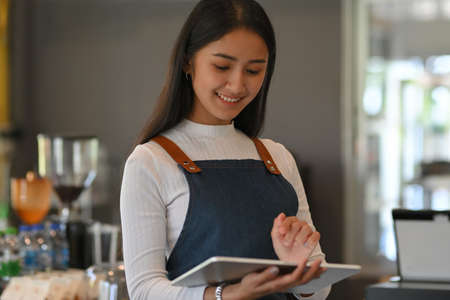 Portrait of female waitress working with digital tablet while standing in front of counter bar in coffee shopの写真素材