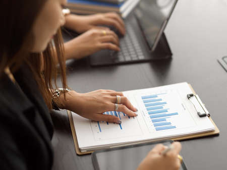 Side view of businesswoman working with business chart on clipboard while sitting in meeting room next to coworkerの写真素材
