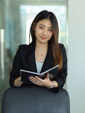 Portrait of female secretary smiling to camera and holding schedule book in office roomの写真素材