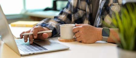 Cropped shot of male hand holding coffee cup and typing on laptop keyboard in home office roomの写真素材