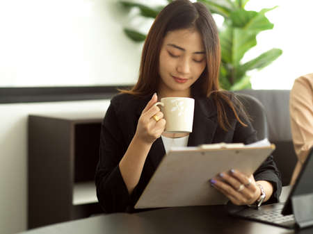 Portrait of businesswoman drinking coffee and reading paperwork on clipboard in meeting roomの写真素材