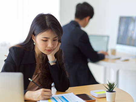 Portrait of businesswoman concentrating on paperwork while working in office room with coworker in backgroundの写真素材