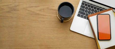 Wooden table with laptop, smartphone, notebooks, coffee cup and coffee cup in home office room