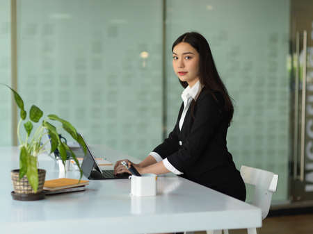 Portrait of businesswoman looking to camera while sitting and working with tablet in office roomの写真素材