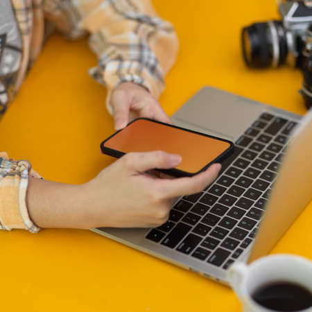 Cropped shot of female photographer working smartphone and laptop on  stylish workspace in home...