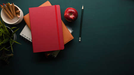 Top view of dark green table with stack of books, pencil, apple, plant pot and copy space, school elementsの写真素材