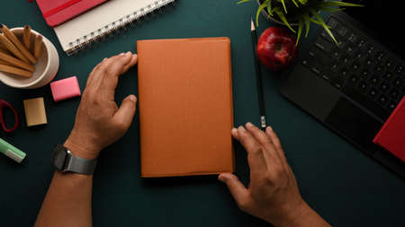 Overhead shot of male hands holding book on dark green study table with stationery and tablet...
