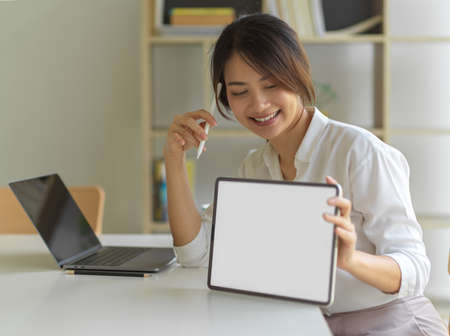 Portrait of female smiling and showing digital tablet with mock up screen on workspace in home office room, clipping pathの写真素材