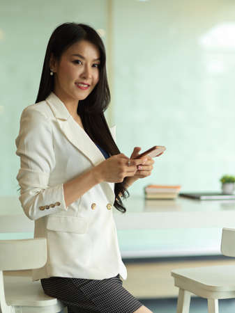 Portrait of businesswoman smiling to camera and using smartphone while standing in office roomの写真素材