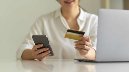 Cropped shot of female online shopping on smartphone and holding credit card while sitting at workspaceの写真素材