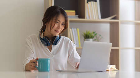 Portrait of female freelancer with headphone holding coffee cup while working with laptop in home office roomの写真素材