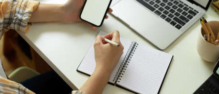 Cropped shot of female freelancer hand writing on blank notebook and using mock up smartphone on workspaceの写真素材