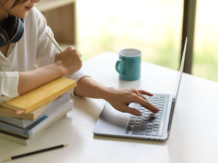 Side view of female university student online studying with laptop and stack of books in living room...