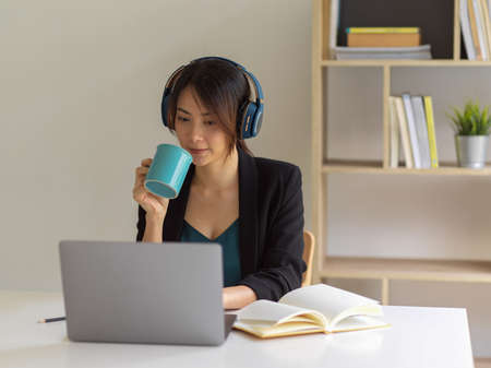 Portrait of businesswoman working from home, using laptop, head phone and drinking coffee in home...