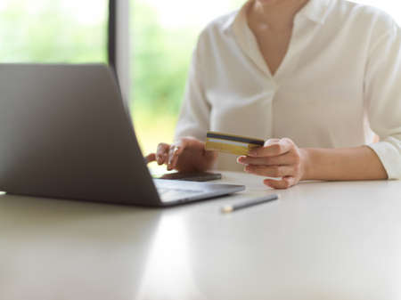 Cropped shot of female hand holding credit card and touching smartphone screen on white desk with...