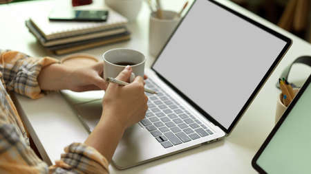 Side view of female hands holding coffee cup while working with laptop on office desk, clipping path