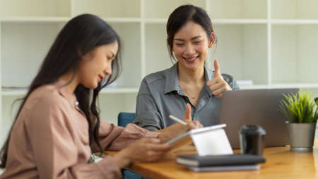 Half-length portraits of female worker giving thumbs up to her colleague while working together in office roomの写真素材