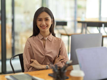 Half-length portrait of young female office worker smiling to camera while sitting at workplaceの写真素材