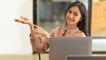 Half-length portrait of female pointing and showing open hand palm with copy space while sitting in office roomの写真素材