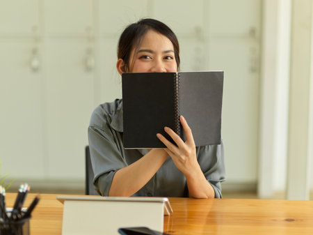 Half-length portrait of young friendly female student hiding her face behind a book while sitting at workplaceの写真素材