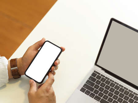 Top view of male hands holding smartphone with mock up screen in his hands on white table with laptop, clipping pathの写真素材