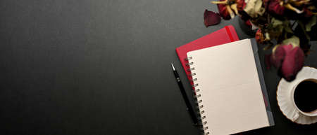 Top view of creative flat lay workspace with notebooks, dried roses, coffee cup and copy space on black tableの写真素材