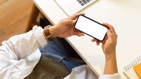 Top view of male freelancer hands holding horizontal smartphone with mock up screen above workspace, clipping pathの写真素材