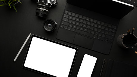 Top view of dark creative flat lay workspace with tablets, smartphone, camera and stationery,...